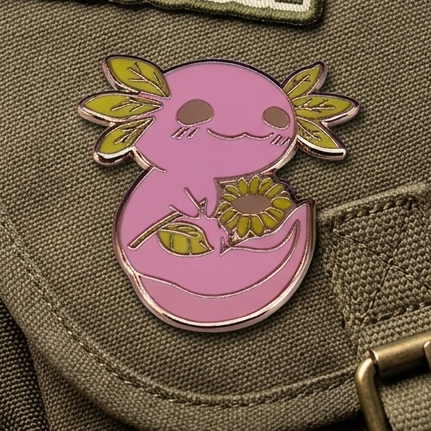 Close-up of an olive green canvas messenger bag with multiple enamel pins and patches attached, including a pink axolotl design