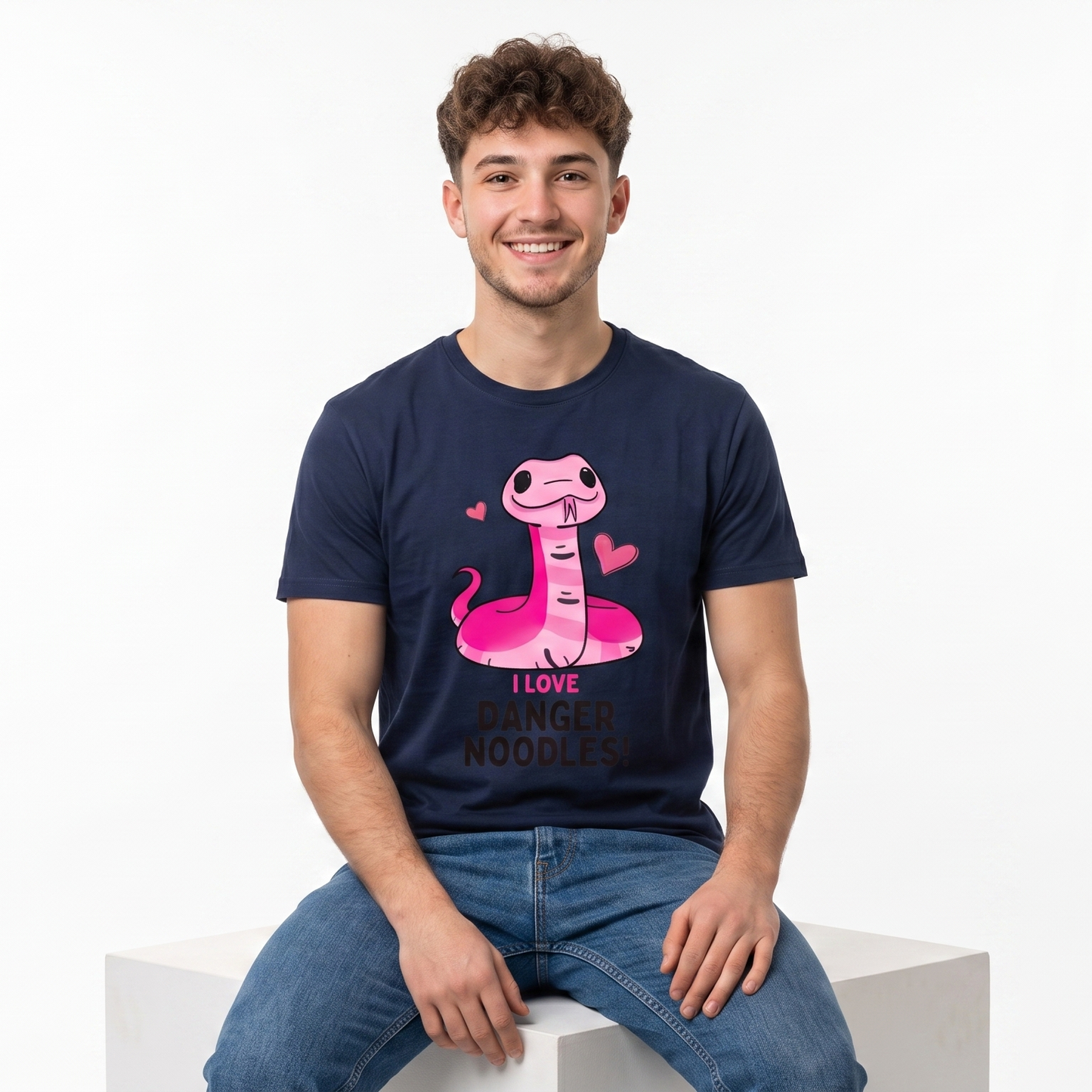 A smiling young man with curly brown hair sits on a white block against a white studio background, wearing a navy blue graphic t-shirt with a pink cartoon snake and the text 'I LOVE DANGER NOODLES' and blue jeans.