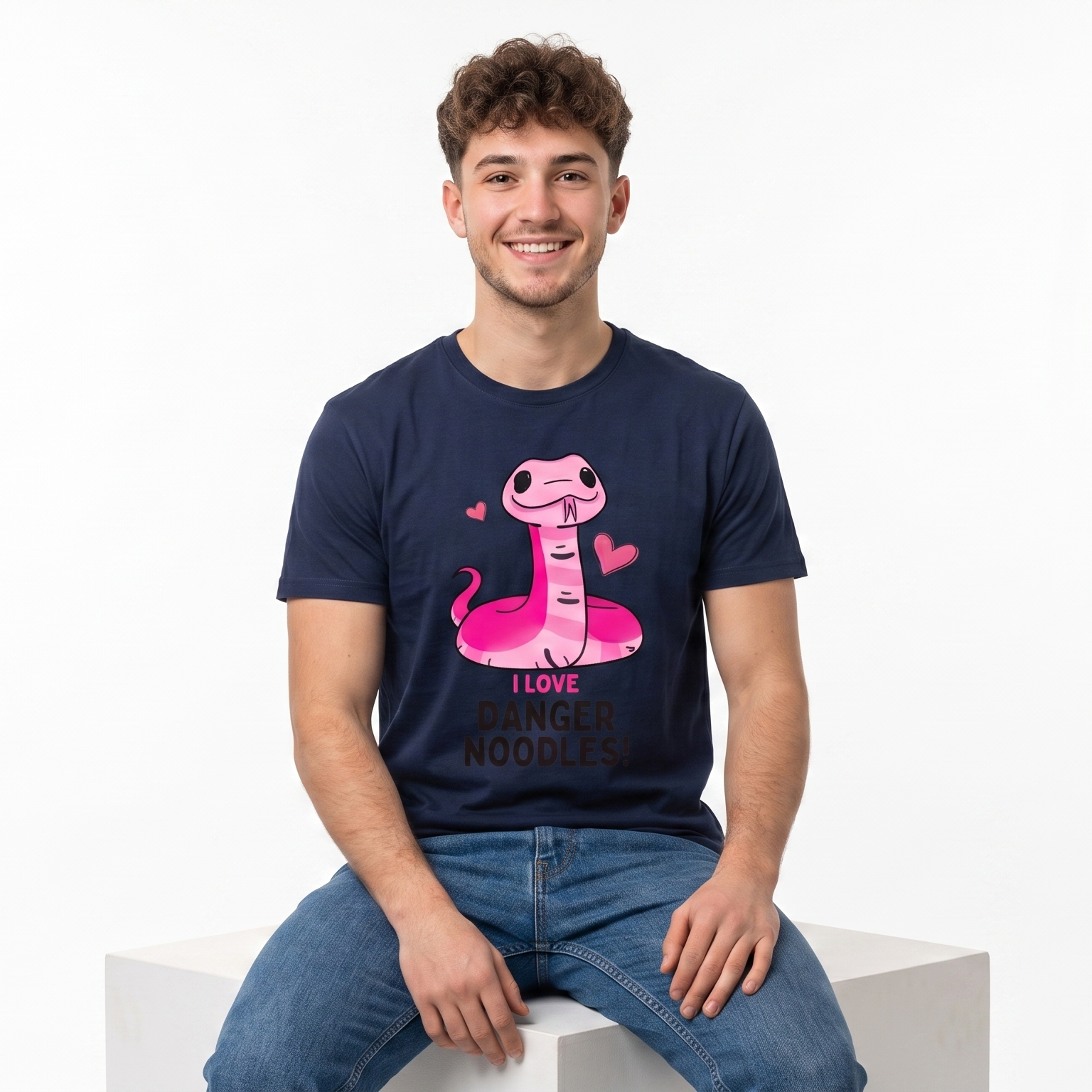 A smiling young man with curly brown hair sits on a white block against a white studio background, wearing a navy blue graphic t-shirt with a pink cartoon snake and the text 'I LOVE DANGER NOODLES' and blue jeans.