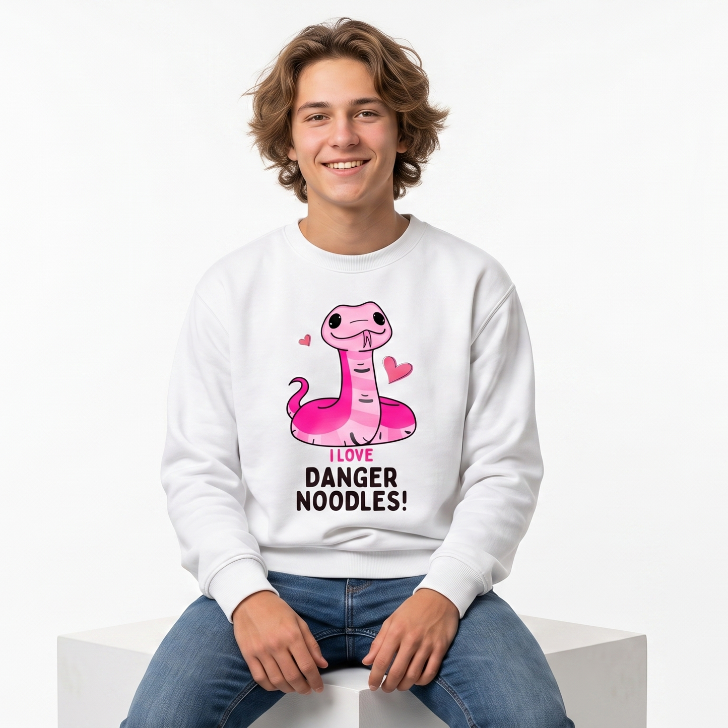 A young man with curly brown hair sits on a white block, wearing a white crewneck sweatshirt with a pink cartoon snake graphic and the text 'DANGER NOODLES!' on the front, paired with blue jeans against a plain white background.