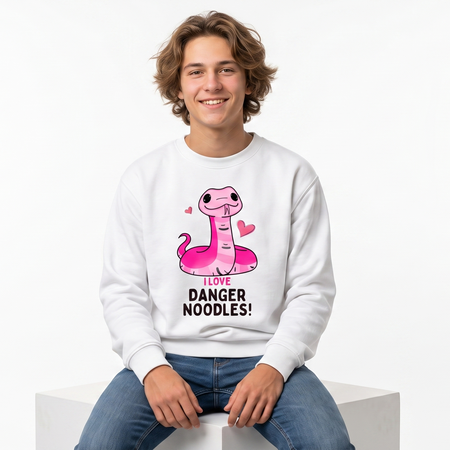 A young man with curly brown hair sits on a white block, wearing a white crewneck sweatshirt with a pink cartoon snake graphic and the text 'DANGER NOODLES!' on the front, paired with blue jeans against a plain white background.