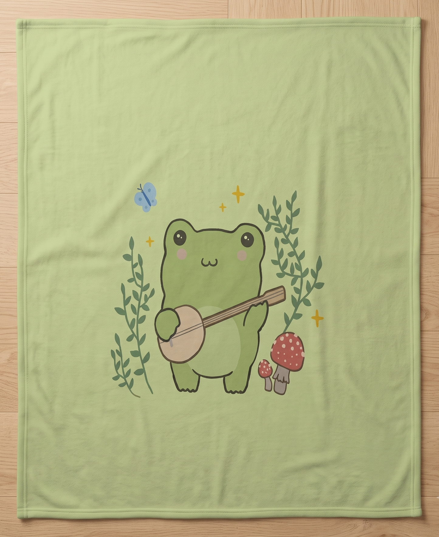 A light green blanket laid flat on a light wood floor, featuring a cute cartoon frog playing a banjo, surrounded by green leaves, red-and-white mushrooms, and a small blue butterfly.