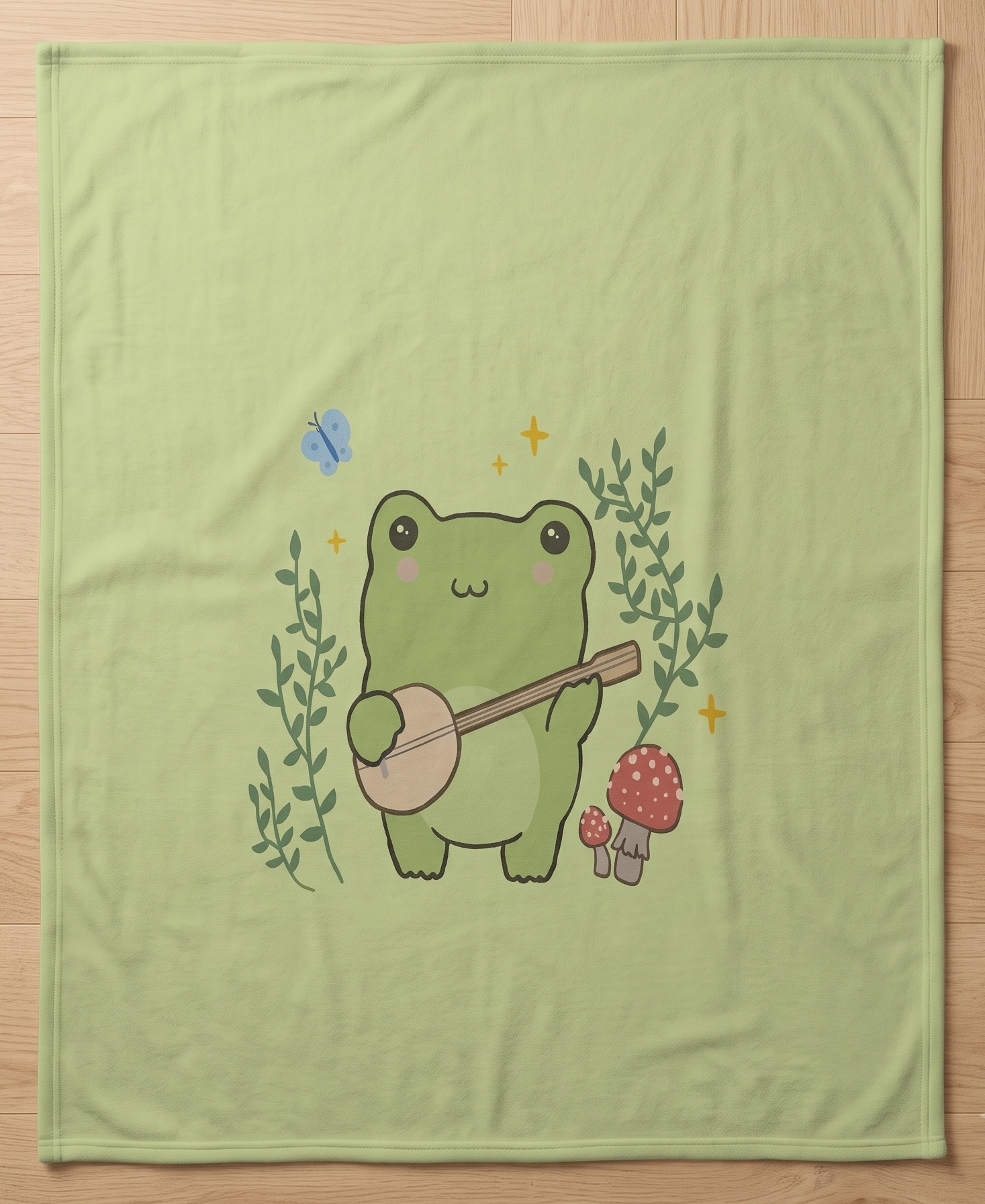 A light green blanket laid flat on a light wood floor, featuring a cute cartoon frog playing a banjo, surrounded by green leaves, red-and-white mushrooms, and a small blue butterfly.