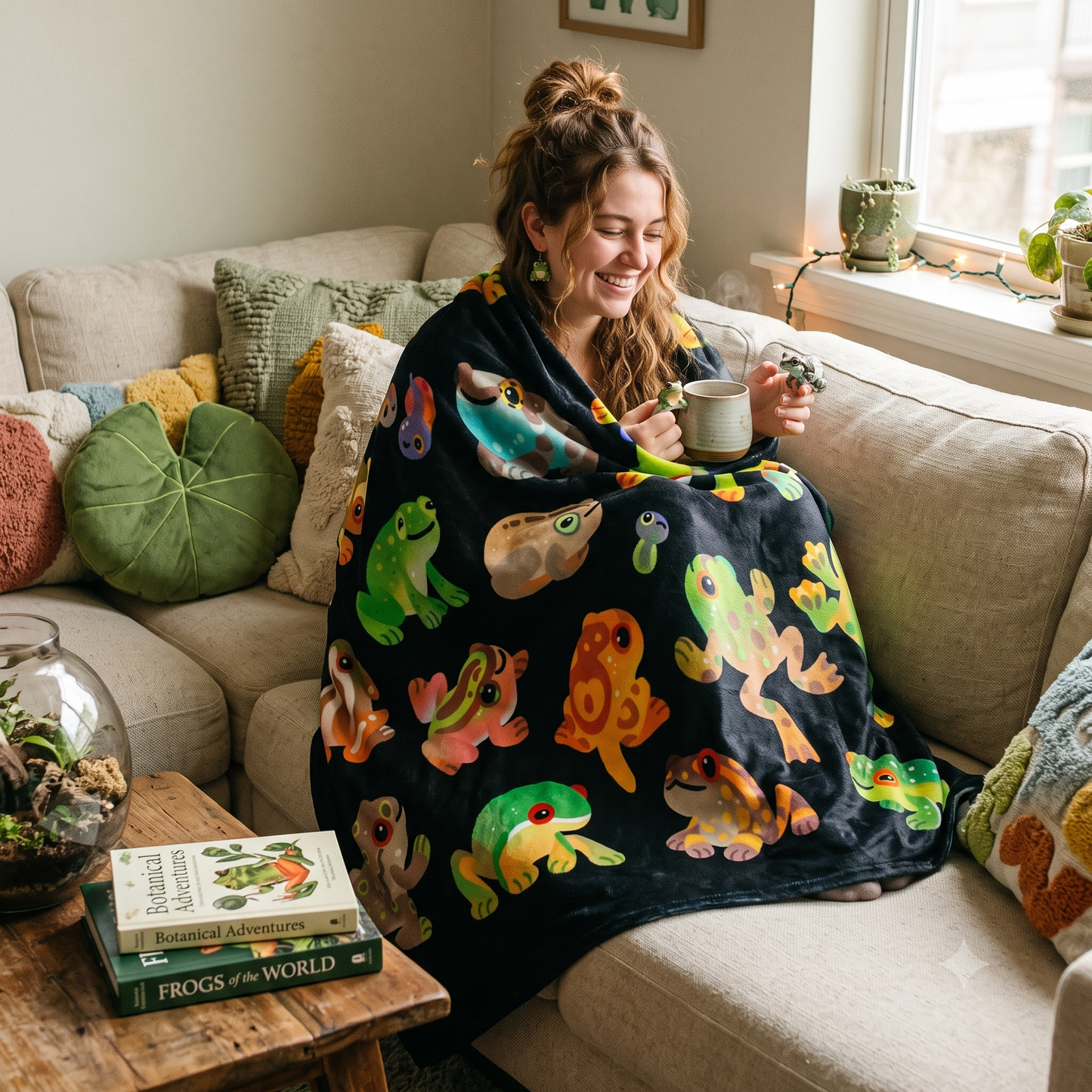 A woman sits on a beige sofa in a living room, wrapped in a dark blanket printed with colorful cartoon frogs. She holds a mug, with books and pillows visible around her.