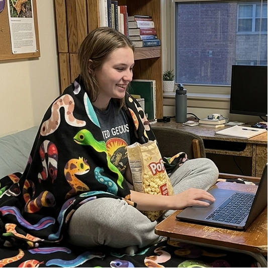 A woman sits on a bed in a dorm room wearing a black blanket with a colorful snake print and holding a bag of chips while using a laptop on a desk.