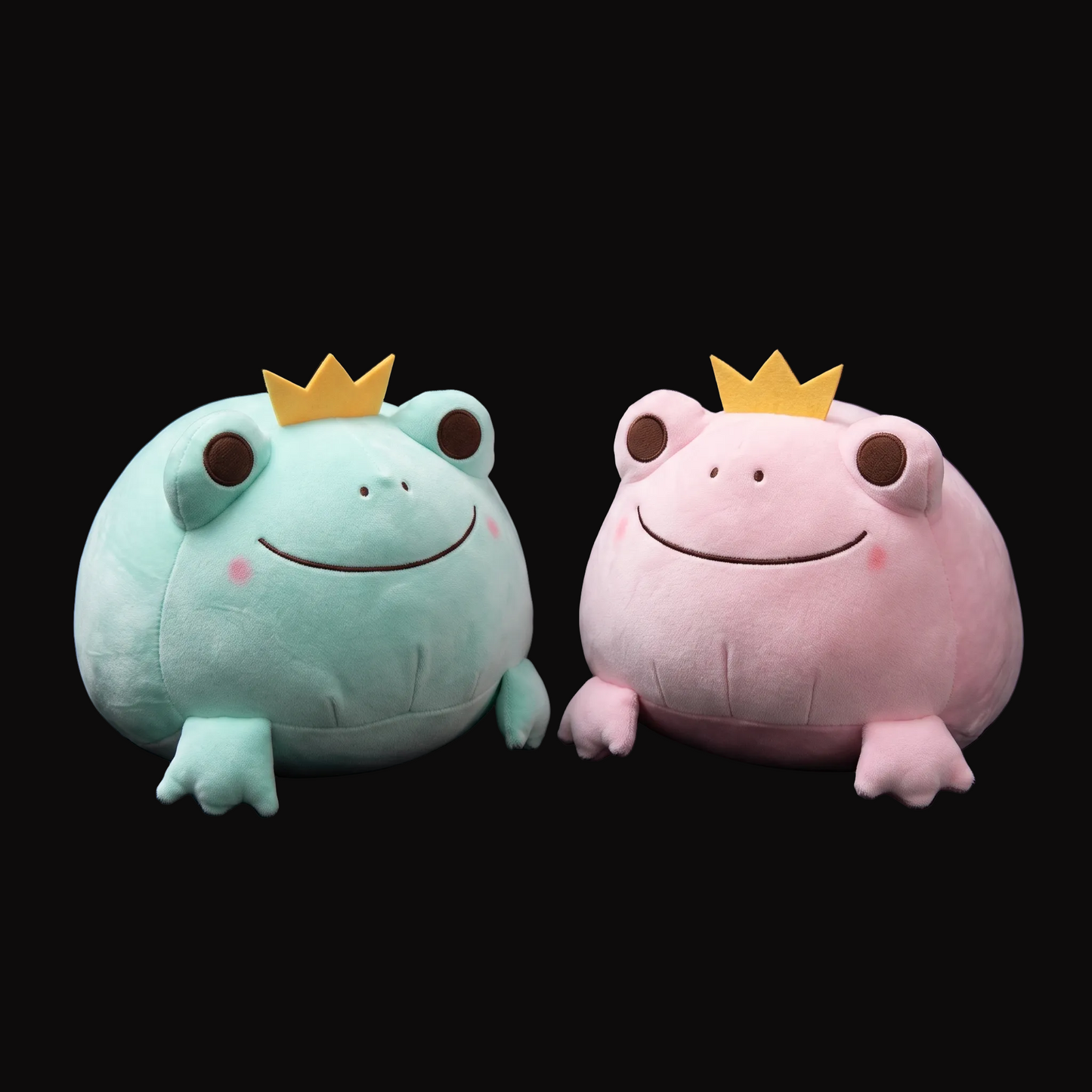 Two round plush frog toys on a black background, one mint green and one light pink, each wearing a small gold crown.