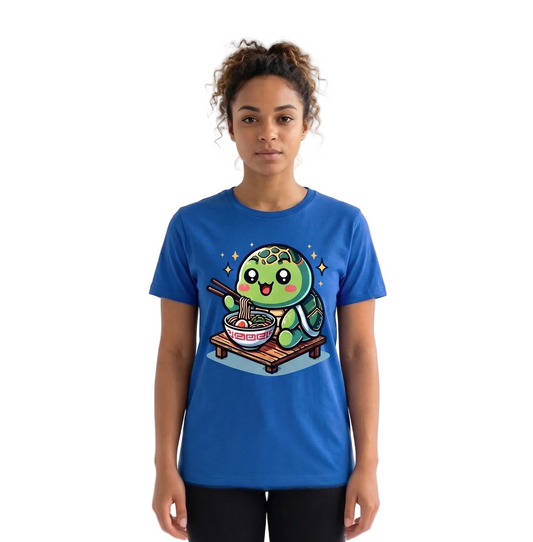 A woman stands against a white studio backdrop wearing a bright blue graphic T-shirt. The shirt features a cartoon green turtle character sitting on a small cart and eating ramen from a bowl with chopsticks, surrounded by small star shapes.