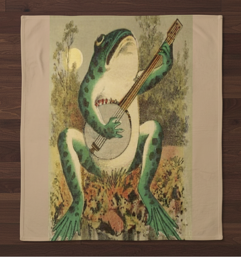A square cream-colored throw blanket featuring a vintage-style illustration of a green frog playing a banjo, laid flat on a dark wood floor.