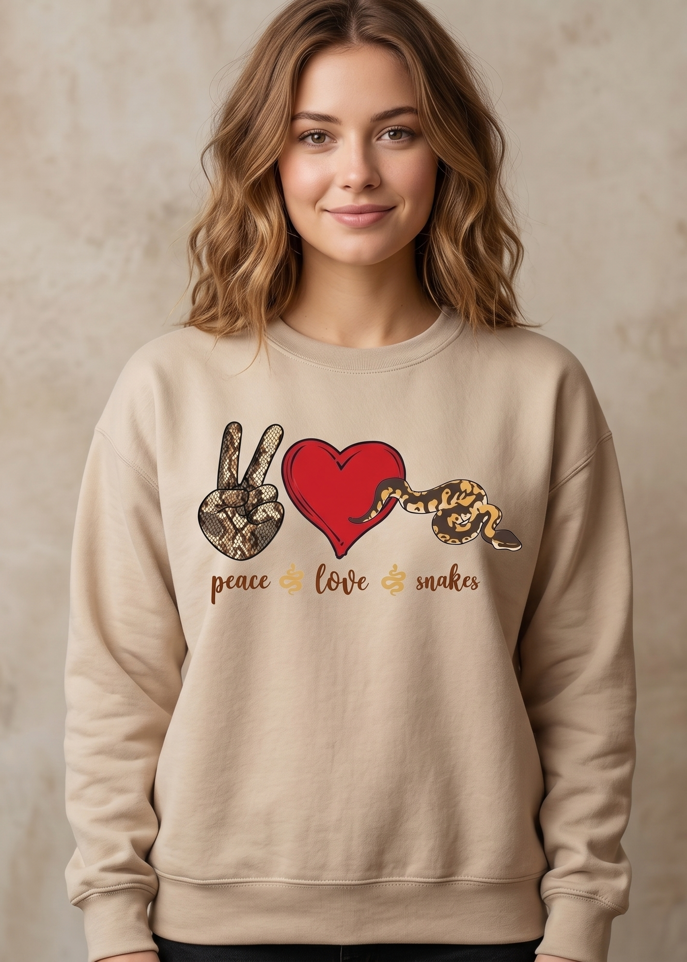 A woman wearing a beige crewneck sweatshirt with a graphic print of a peace hand sign, a red heart, a snake, and the words 'peace love snakes' in gold, set against a textured beige background.