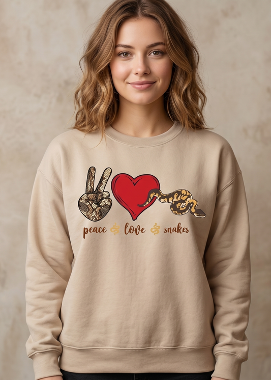 A woman wearing a beige crewneck sweatshirt with a graphic print of a peace hand sign, a red heart, a snake, and the words 'peace love snakes' in gold, set against a textured beige background.