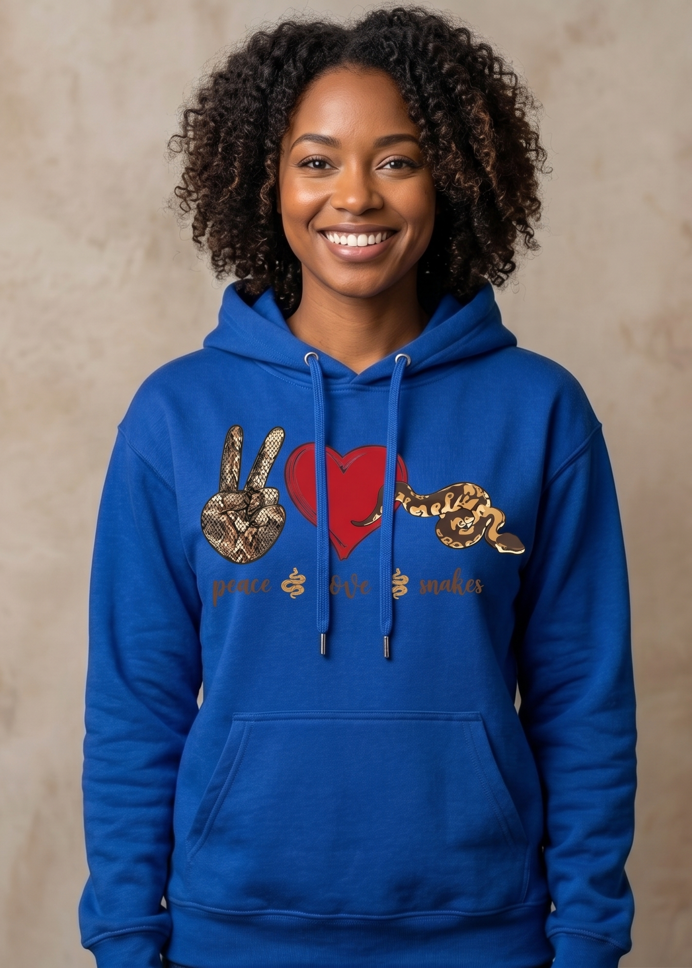 A smiling woman with curly hair wearing a bright blue hoodie with a graphic on the chest featuring a red heart, peace sign, snake, and the words 'peace love makes', posed against a textured beige background.