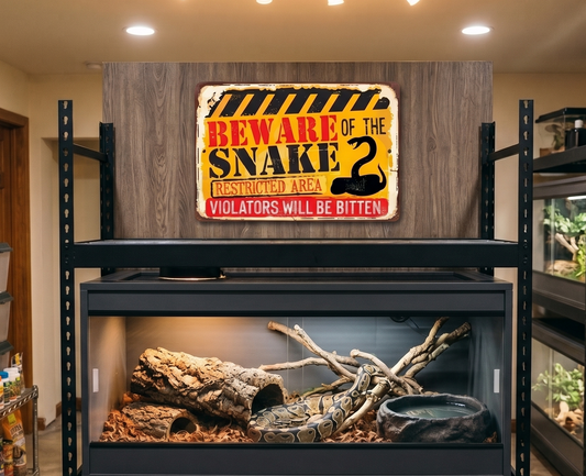 A large black metal reptile rack holding a glass terrarium with wood logs and a bowl, set in a home reptile room. A decorative sign reading 'Beware of the Snake' hangs above the rack, with shelving and other enclosures visible in the background.