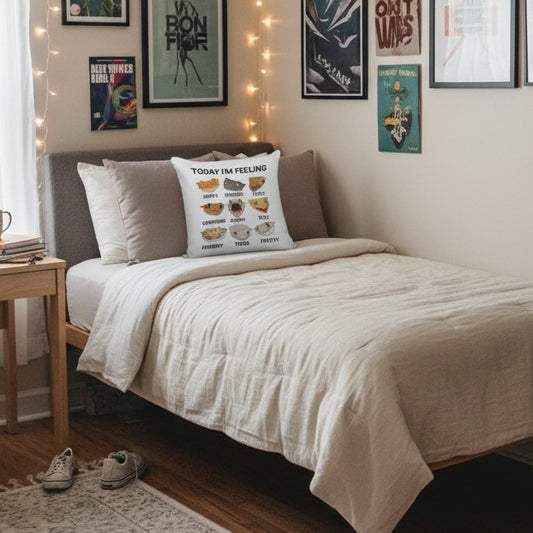 A square white throw pillow with a printed cartoon gecko chart reading 'TODAY I'M FEELING HAPPY FOCUSED CRAZY CONFUSED ANGRY SILLY HUNGRY TIRED PRETTY' is styled on a bed with gray and white pillows.