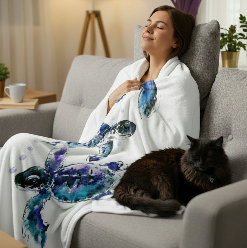 A woman relaxes in a gray armchair wrapped in a white throw blanket featuring a blue and purple watercolor sea turtle design. A dark cat rests on the armchair beside her, with a softly lit living room in the background.